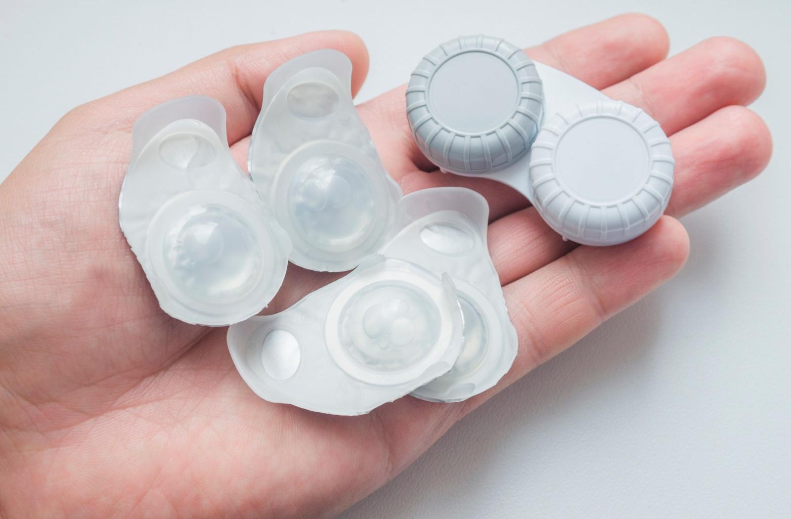 A hand holding unopened contact lens blister packs and a contact lens case.